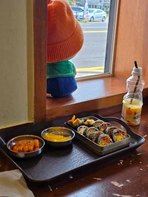 창가 테이블 위 검은 트레이에 담긴 김밥과 단무지, 떡볶이 소스