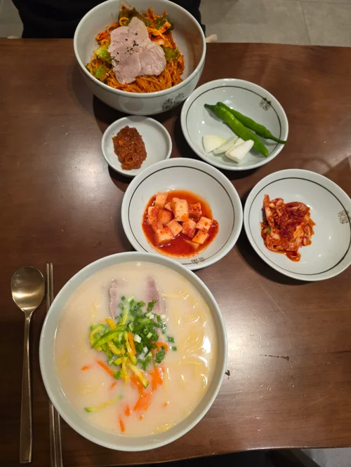 테이블 위에 비빔국수와 떡국, 김치 등 다양한 반찬이 차려진 한식 정식