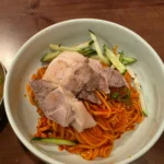 흰 그릇에 담긴 매콤한 비빔냉면 위에 수육과 오이채가 올려진 모습