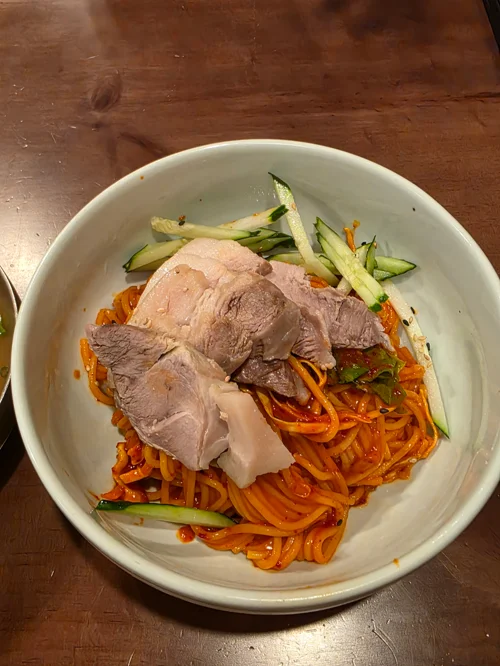 흰 그릇에 담긴 매콤한 비빔냉면 위에 수육과 오이채가 올려진 모습