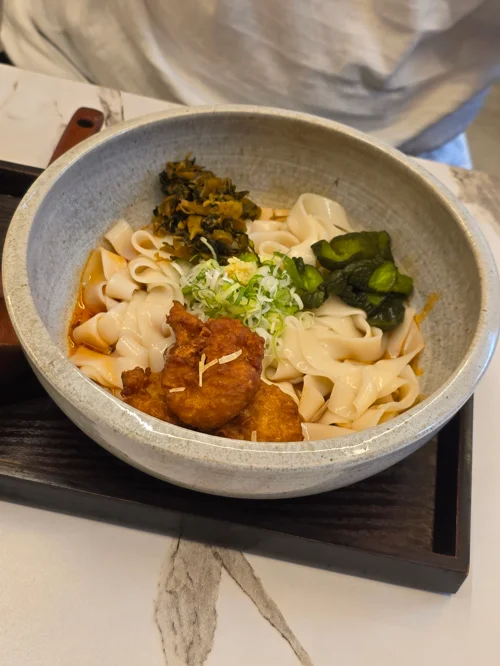 회색 그릇에 담긴 칼국수 위에 튀긴 닭고기와 채소 토핑