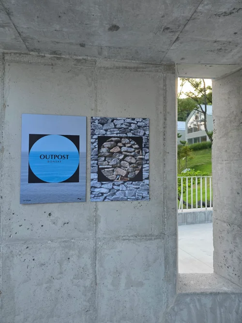 콘크리트 벽에 걸린 OUTPOST 로고와 돌담 작품, 창밖으로 보이는 녹색 풍경