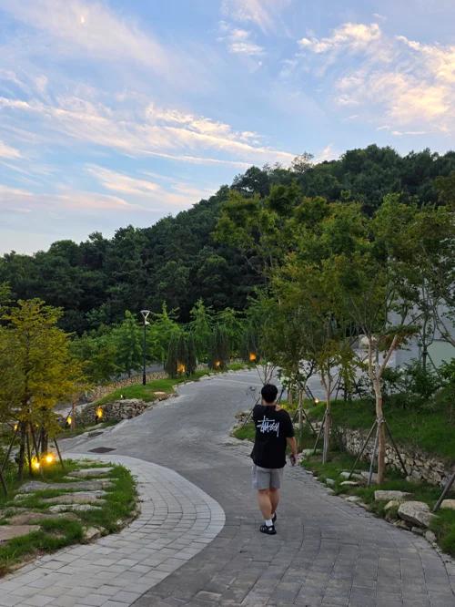 곡선형 산책로를 따라 걷는 사람과 양쪽 조명, 배경의 숲과 저녁 하늘