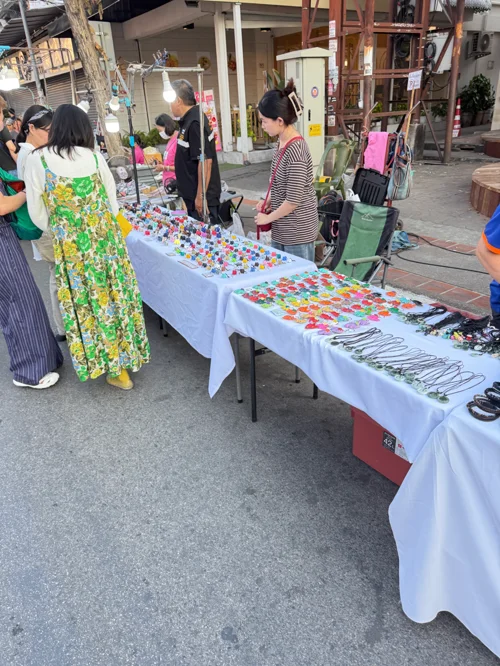 야외 테이블에 진열된 다양한 색상의 수제 액세서리를 둘러보는 사람들