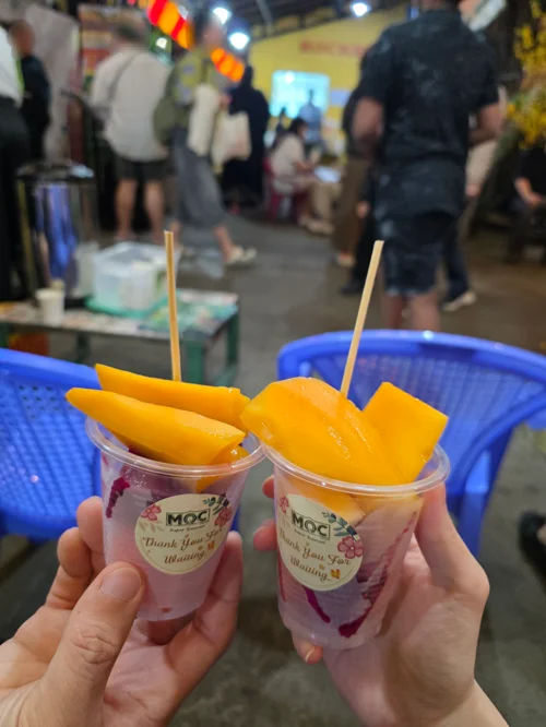 두 손이 망고 조각이 담긴 플라스틱 컵을 들고 있는 야시장 풍경