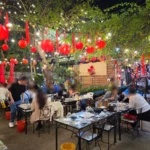 빨간 등롱과 전구 조명이 매달린 나무 아래 손님들이 식사하는 가든 레스토랑 내부