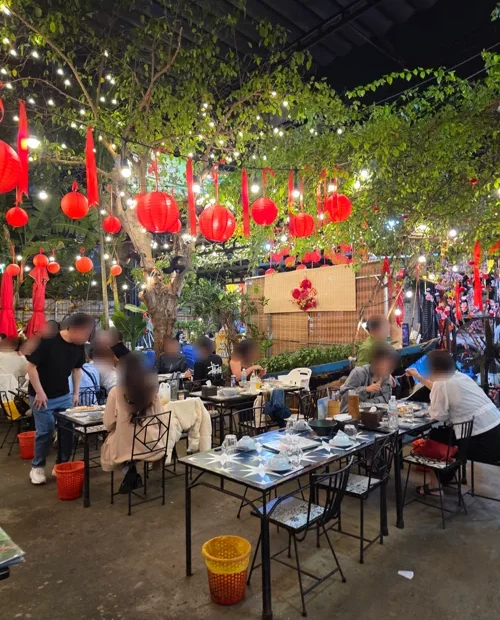 빨간 등롱과 전구 조명이 매달린 나무 아래 손님들이 식사하는 가든 레스토랑 내부