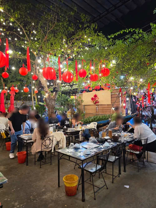 빨간 등롱과 전구 조명이 매달린 나무 아래 손님들이 식사하는 가든 레스토랑 내부