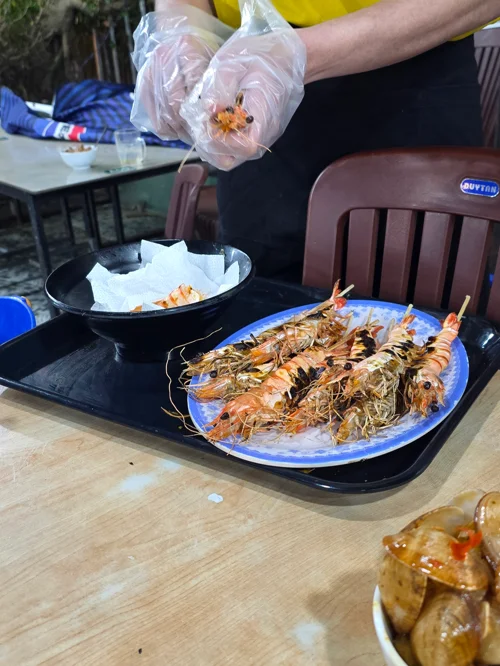 야외 테이블 위 파란 접시에 담긴 구운 새우와 손질 중인 모습