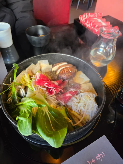 김이 모락모락 나는 스키야키 냄비 - 청경채, 두부, 버섯, 소고기, 우동면이 끓고 있는 모습