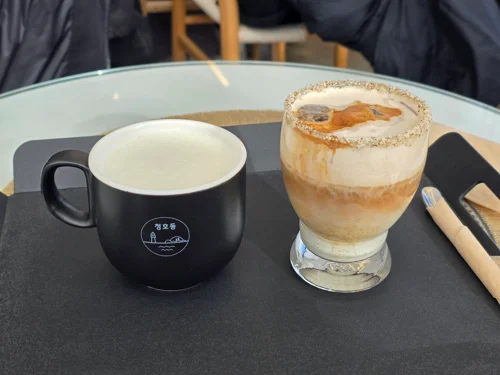 검은 머그컵과 레이어드 음료가 담긴 투명 잔이 카페 테이블에 놓여있음