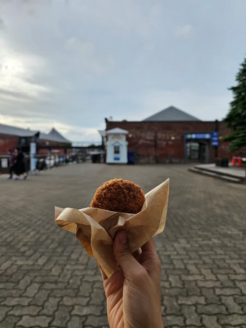 붉은 벽돌 건물 앞 광장에서 손에 든 황금빛 고로케