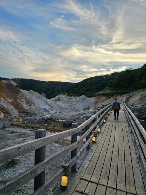 저녁 등불이 켜진 나무 데크 산책로를 걷는 사람과 유황 지열 지대가 펼쳐진 노보리베츠 지옥계곡 풍경