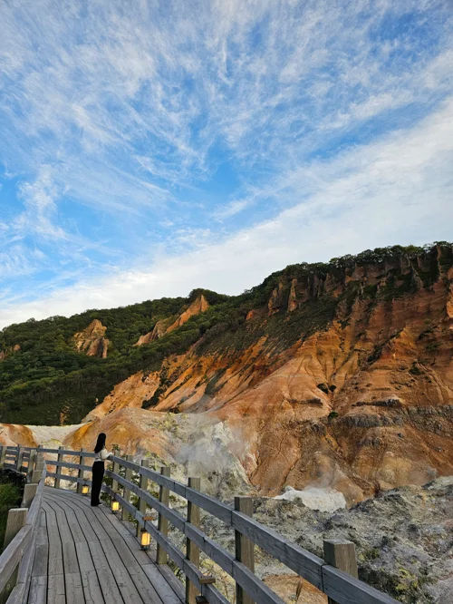 붉은 화산 절벽과 유황 연기가 피어오르는 지고쿠다니 데크 위에 서 있는 여행자
