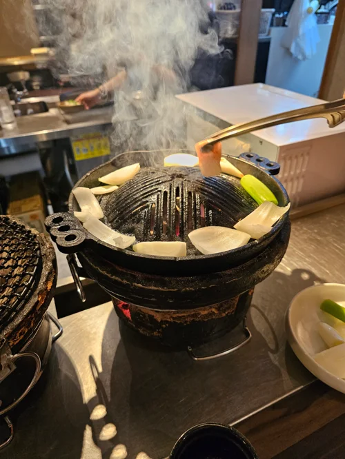 연기가 피어오르는 철제 숯불 그릴 위에 마늘과 양파가 구워지는 모습