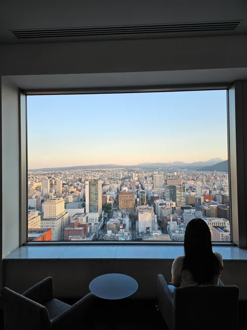 고층 라운지 창가에서 삿포로 도시 전경과 석양을 바라보는 여성의 실루엣
