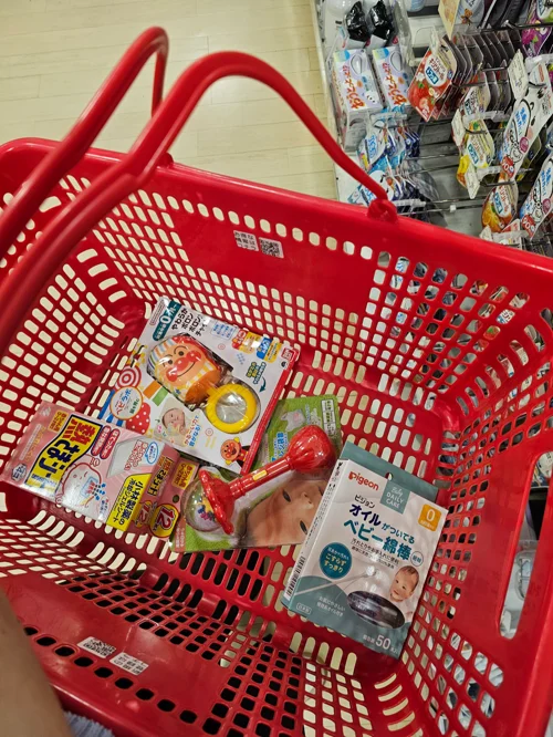일본 마트의 빨간 쇼핑 바구니에 담긴 아기 장난감과 피존 면봉 등 유아용품