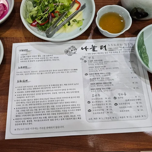 나눌터 도토리오리 전문점 메뉴판이 반찬 그릇들과 함께 테이블에 놓인 모습