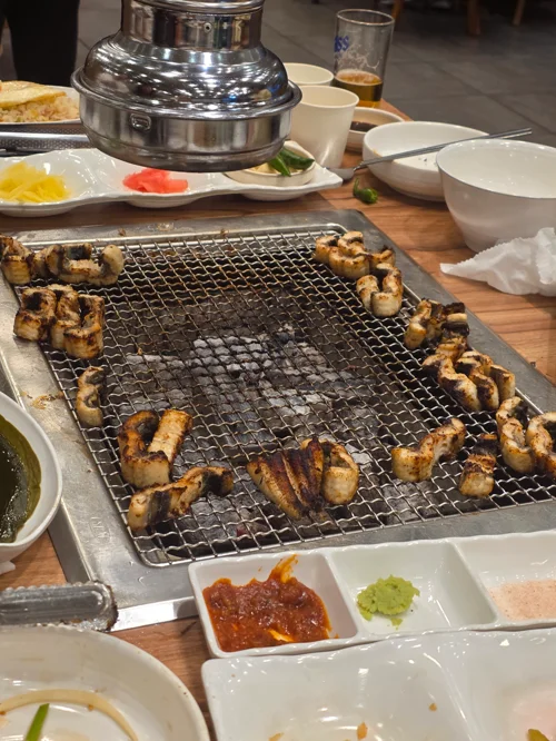 숯불 철망 그릴 위에 노릇하게 구워진 장어 조각들과 소스가 놓인 장어구이 식당 테이블