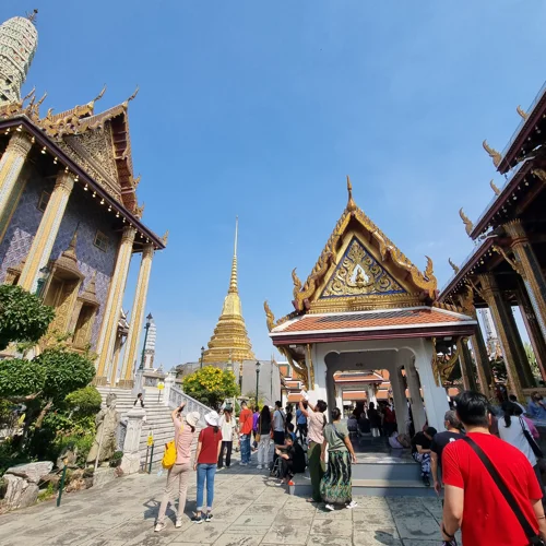 방콕 왓 프라깨우 경내에서 황금 첨탑과 전통 사원 건물을 배경으로 관광객들이 걷고 있는 모습