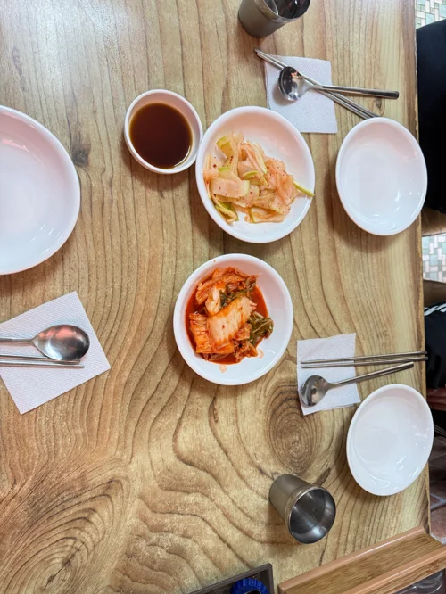 나무 테이블 위에 김치와 무김치 반찬, 수저가 세팅된 한식당 테이블