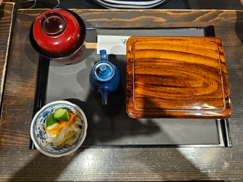 검은 쟁반 위에 붉은 국그릇, 파란 도자기 주전자, 나무 도시락 상자, 절임 채소 반찬이 놓인 일식 세트